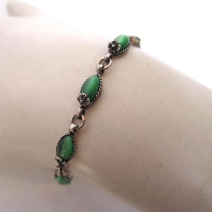 May include: A silver-toned bracelet featuring oval, green gemstones. The bracelet has a delicate design with small floral accents between each gemstone. The gemstones have a smooth, polished appearance, and the bracelet is displayed on a white surface.