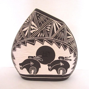 May include: Black and white ceramic vase with a distinctive shape. The vase showcases a sun and two bear figures with geometric designs. The top of the vase has a detailed pattern. The vase is a decorative item.