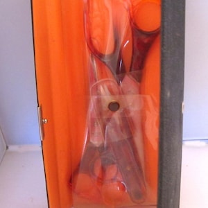 May include: A black case with an orange interior contains various scissors and cutting implements. The tools are packaged in clear plastic. The case is open, displaying the contents. The scissors have orange handles and dark blades.