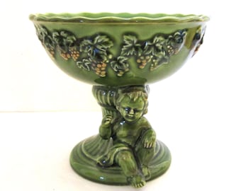 May include: Green ceramic pedestal bowl with a cherub base. The bowl features embossed grape and leaf designs around the rim. The cherub is seated on a round base, supporting the bowl above. A decorative piece.