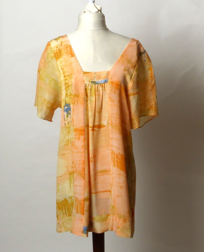 Puede incluir: Un vestido corto de manga corta, holgado, con cuello redondo y fondo de color melocot&oacute;n. El vestido tiene un patr&oacute;n geom&eacute;trico en tonos de naranja, amarillo y azul.