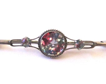 May include: A silver brooch with a large, round, multi-colored gemstone surrounded by a silver bezel. Two smaller, round, multi-colored gemstones are on either side of the large gemstone.