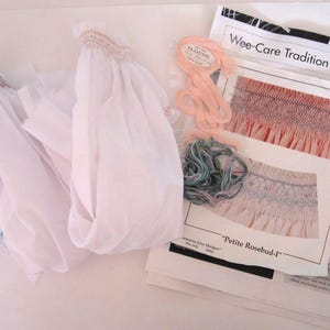 Può includere: Un kit da cucito con pezzi di tessuto bianco con arricciature a punto smock sulla parte superiore, matasse di filo da ricamo color pesca e blu variegato, e un libretto di istruzioni intitolato Wee-Care Tradition Petite Rosebud-I di Ruth Engle.