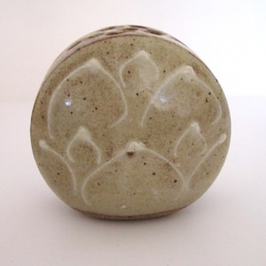 Puede incluir: Objeto cerámico redondo y moteado, de color verde-marrón claro. La superficie presenta un patrón repetido de diseños florales estilizados. El borde superior tiene pequeños agujeros espaciados. El diseño es sencillo y orgánico.