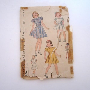 Op de afbeelding: Een vintage Simplicity patroon envelop, nummer 3234, maat 4, met illustraties van kinderen in jurken met pofmouwen en uitlopende rokken. De papieren envelop is verouderd, verkleurd en vertoont aanzienlijke slijtage en scheuren.