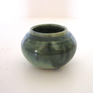 May include: A small, round ceramic pot with a glossy finish in shades of deep forest green and lighter sage green. The vessel features a wide, bulbous body that tapers toward a circular opening at the top, showcasing smooth, blended color transitions.