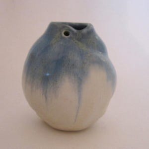 Pode incluir: Um pequeno vaso de cerâmica feito à mão com um corpo arredondado e um pescoço estreito. O vaso é principalmente branco, com uma cobertura azul que escorre pelas laterais. Possui um pequeno orifício perto do topo.