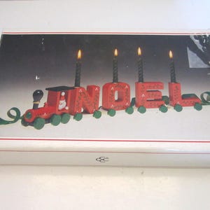 Può includere: Una scatola di cartone rettangolare con l'immagine di un portacandele a forma di trenino in legno rosso che compone la parola NOEL. Il treno ha ruote verdi, una figura di Babbo Natale nella locomotiva e quattro candele nere nelle lettere.