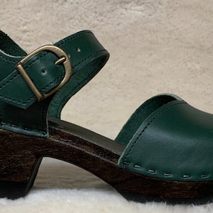 May include: Dark green leather clog with a wooden heel and an adjustable ankle strap with a gold buckle.