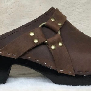 May include: Brown leather clog with a black wooden heel and a buckle strap with metal accents.