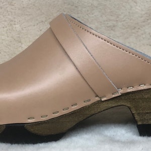 May include: A pair of beige leather clogs with a wooden heel and a strap across the top. The clogs have a stitched seam around the edge of the sole.
