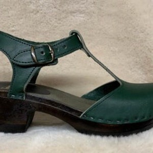 May include: Dark green leather heeled clogs with a t-strap and buckle closure. The clogs have a dark brown wooden sole and heel.