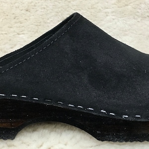 May include: Black suede clogs with a wooden platform sole. The clogs have a stitched seam around the edge of the sole and the brand name "Gveni" is visible on the insole.