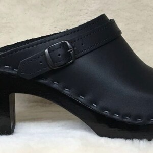 May include: Black leather heeled clog with a buckle strap and a stitched seam around the sole.