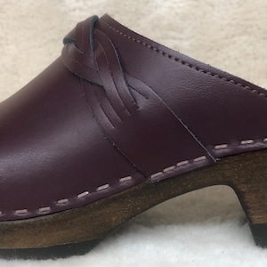 May include: Burgundy leather clog with a braided strap detail and a wooden heel.