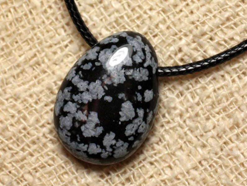 May include: Black and white snowflake obsidian pendant on a black cord. The pendant is a smooth, oval shape with a speckled pattern.