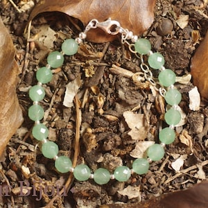 Pode incluir: Uma pulseira de contas verde claro com fecho prateado. A pulseira é composta por contas redondas e facetadas, enfiadas com uma corrente prateada. A pulseira é exibida sobre uma cama de folhas e galhos marrons.