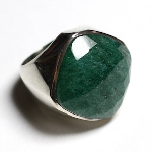 Pode incluir: Um anel de prata com uma grande pedra preciosa verde esmeralda facetada. A pedra tem um corte geométrico, que reflete a luz. A banda do anel é de prata polida, criando contraste com a pedra verde escura. Em fundo branco.