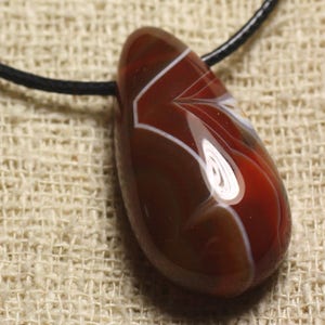May include: A teardrop-shaped pendant made of red agate with white and brown swirling patterns. The pendant is attached to a black cord.