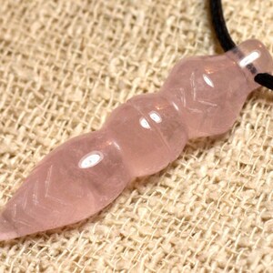 Peut inclure: Un collier avec un pendentif en quartz rose. Le pendentif rose pâle est sculpté dans une forme unique avec une extrémité pointue et des sections arrondies. Il est suspendu à un cordon noir. Le fond est un tissu beige texturé.