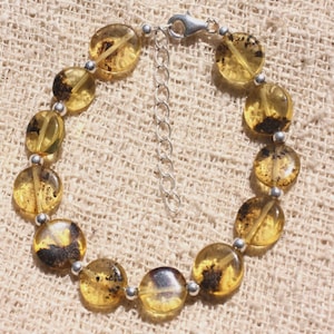 Puede incluir: Un brazalete hecho de cuentas redondas de color ámbar con inclusiones negras. Las cuentas están enhebradas en una cadena de plata.