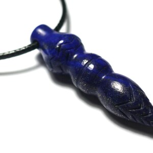 Peut inclure: Pendentif en lapis-lazuli bleu foncé avec un motif sculpté, suspendu à un cordon noir. Le pendentif a une forme cylindrique avec une surface texturée et des gravures complexes. Le fond est blanc.