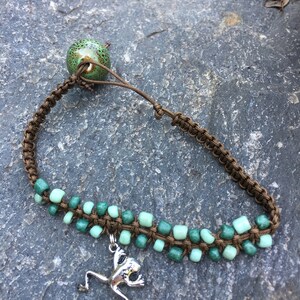 May include: A brown cord bracelet with green and light green beads and a silver frog charm. The bracelet has a green and brown speckled bead at the end.