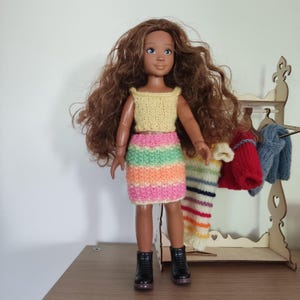 May include: A doll wearing a yellow knitted crop top and a striped skirt in pink, green, orange, and white. The doll has long brown hair and black boots. A wooden clothes rack with miniature knitted garments is in the background.