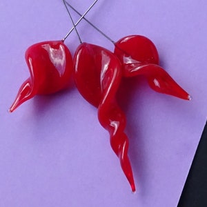 Peut inclure: Trois pièces d'art en verre rouge vif faites à la main, de forme torsadée et organique. Chaque pièce est fixée à un fin fil d'argent, suggérant qu'il s'agit d'éléments d'un design ou d'un bijou plus large. Le fond est violet clair.
