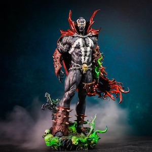 May include: A detailed figurine of the comic book character Spawn. The figure is predominantly black and gray, with a red cape and accents. It stands on a base with skulls and a cross, with green and red accents.