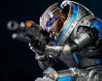 May include: A detailed figurine of a science fiction character in a futuristic armored suit, holding a rifle. The armor is primarily metallic gray with blue accents. The character's head is visible, with brown skin and blue markings.