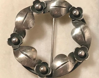 May include: A silver brooch in the shape of a wreath with five stylized flowers and leaves.