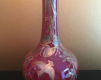 May include: A red glass vase with a silver and gold floral design. The vase has a long neck and a round body.