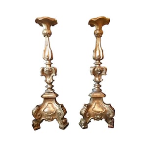 May include: Two antique gold-colored candlesticks with ornate, detailed designs. Each candlestick features a wide, decorative base, a slender, detailed stem, and a flared top. The candlesticks are made of a metallic material and have a vintage aesthetic.
