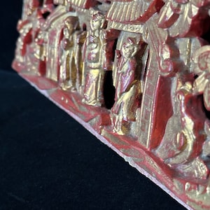 May include: A carved wooden panel with a red and gold paint finish. The panel depicts a scene with multiple figures and buildings. The figures are dressed in traditional clothing and the buildings have ornate roofs.