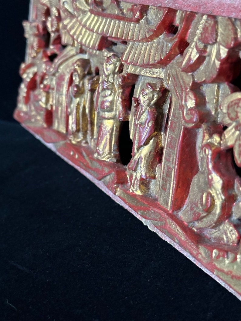 May include: A carved wooden panel with a red and gold paint finish. The panel depicts a scene with multiple figures and buildings. The figures are dressed in traditional clothing and the buildings have ornate roofs.