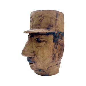 May include: A ceramic sculpture of a person's head wearing a brown cap with a star on it. The sculpture is made of brown clay and has a textured surface.