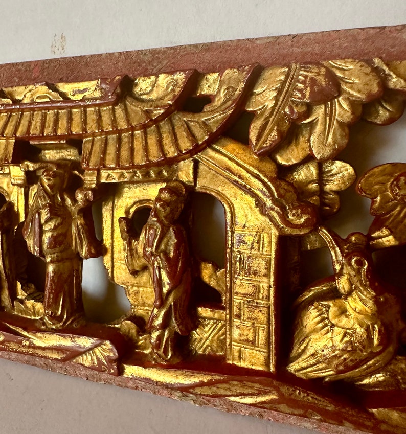 May include: A carved wooden panel with a gold and red finish. The panel depicts a scene with figures, a building, and a bird. The panel is a traditional Chinese art form.