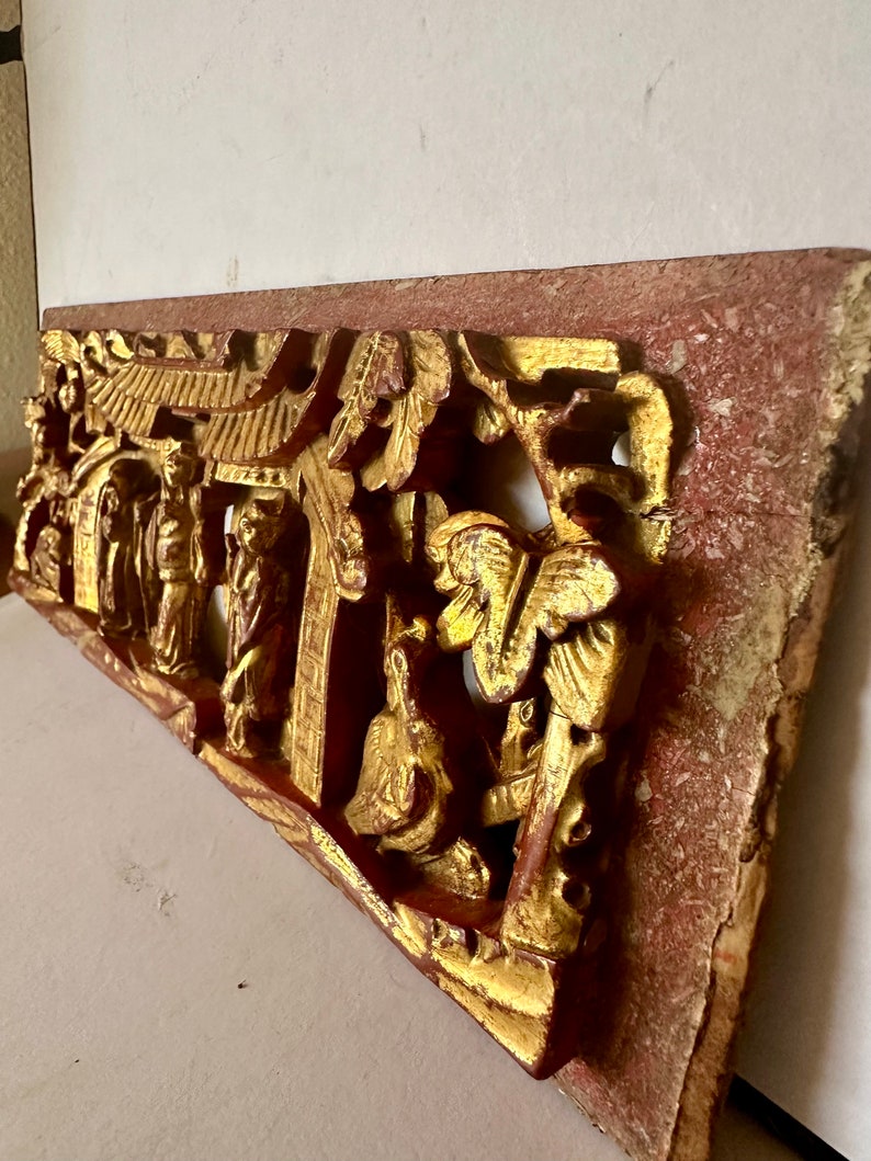 May include: A carved wooden panel with a red and gold finish. The panel depicts a scene with figures and a pagoda. The panel is intricately carved and has a distressed look.