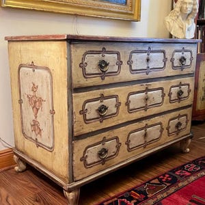 May include: Antique chest of drawers with a cream-colored finish and decorative painted accents. The chest features three drawers with ornate hardware and a painted floral design on the side. The chest is supported by four curved legs.