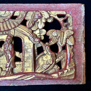 May include: A carved wooden panel with intricate details. The panel is painted in shades of red and gold, and features a scene of two figures in a garden setting. The figures are dressed in traditional Chinese clothing, and the garden is filled with flowers and trees.