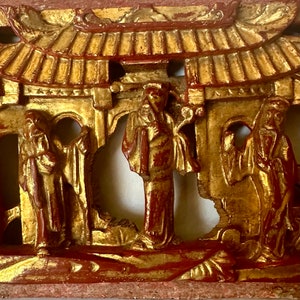 May include: A carved wooden panel with a gold and red paint finish. The panel depicts a scene with three figures in traditional Chinese clothing standing in front of a building with a roof. The figures are carved in relief and the building is carved in openwork.