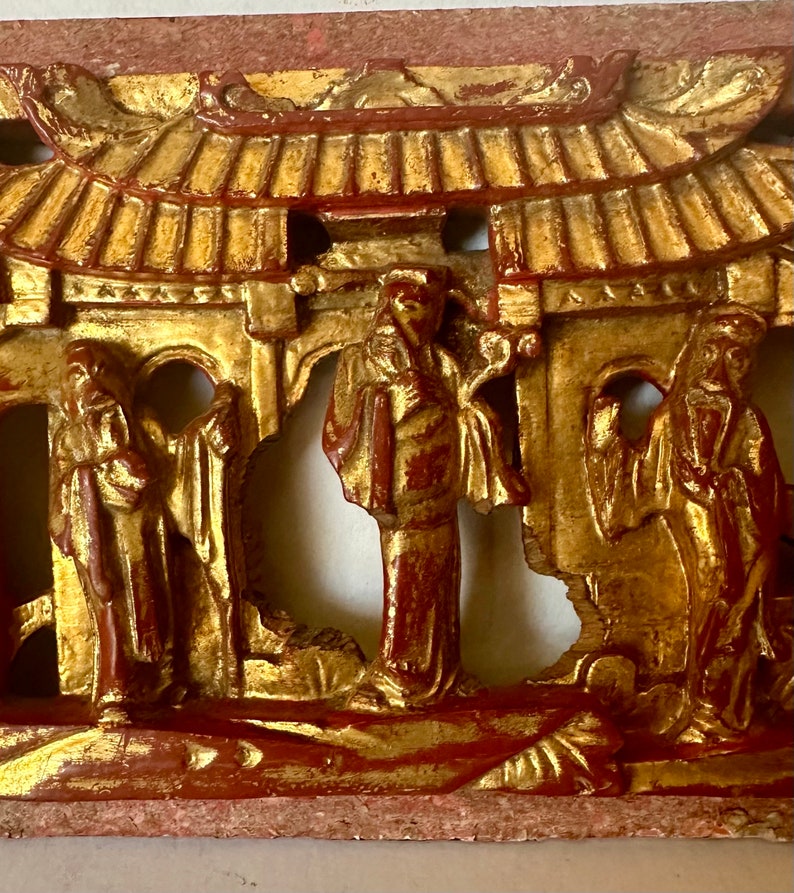 May include: A carved wooden panel with a gold and red paint finish. The panel depicts a scene with three figures in traditional Chinese clothing standing in front of a building with a roof. The figures are carved in relief and the building is carved in openwork.
