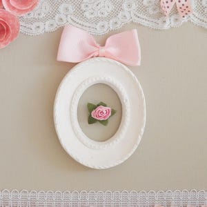 May include: A white oval picture frame with a pink bow and a small pink rose inside. The frame is decorated with a textured border. Pink paper roses and lace trim the top and bottom of the image.