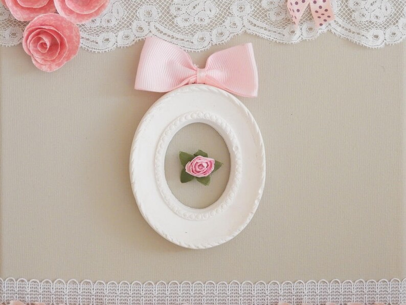 May include: A white oval picture frame with a pink bow and a small pink rose inside. The frame is decorated with a textured border. Pink paper roses and lace trim the top and bottom of the image.