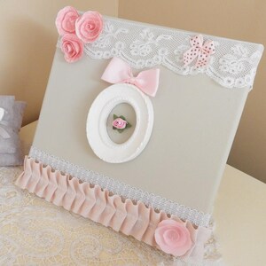 May include: A decorative canvas with a light gray background, adorned with pink fabric roses, a pink bow, and a white oval frame. The canvas is trimmed with white lace and a pink ruffled fabric. A small gray pillow with a white butterfly is visible.