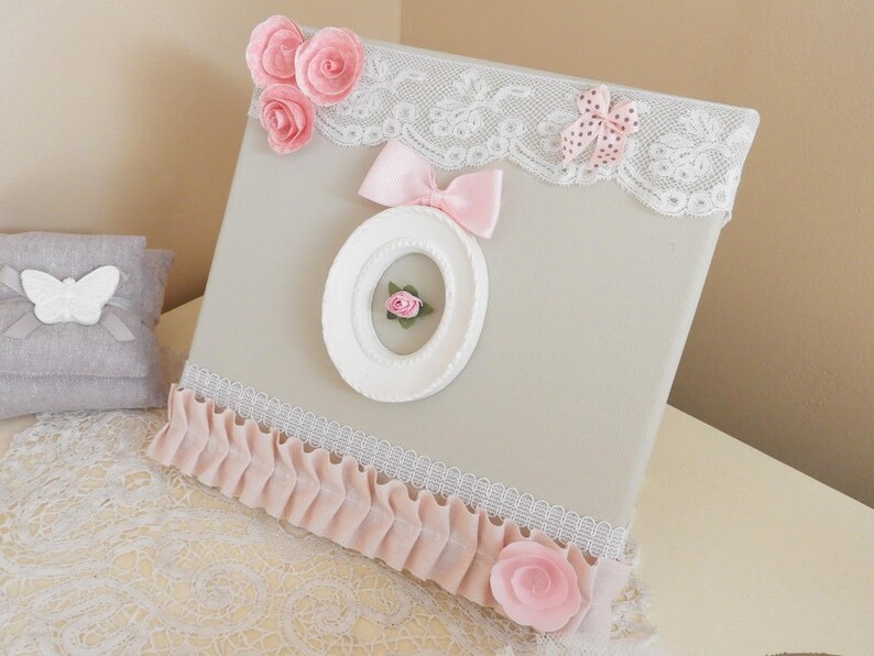 May include: A decorative canvas with a light gray background, adorned with pink fabric roses, a pink bow, and a white oval frame. The canvas is trimmed with white lace and a pink ruffled fabric. A small gray pillow with a white butterfly is visible.