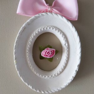May include: A white, oval-shaped frame with a pink ribbon bow at the top. Inside the frame is a small pink rose with green leaves. The frame has a beaded inner border and a textured outer edge. Decorative lace is visible.