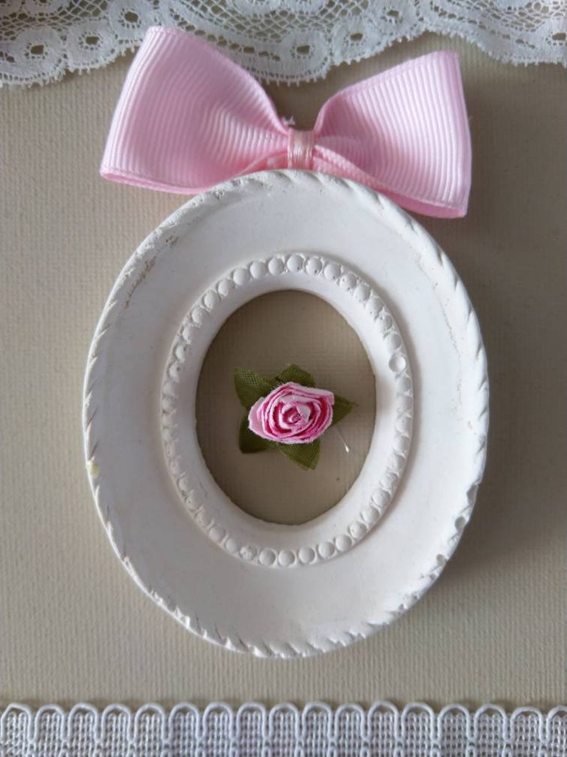 May include: A white, oval-shaped frame with a pink ribbon bow at the top. Inside the frame is a small pink rose with green leaves. The frame has a beaded inner border and a textured outer edge. Decorative lace is visible.