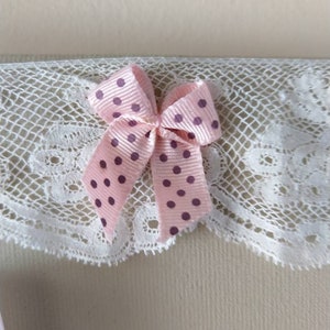 May include: A close-up of a pink polka-dot bow with a white lace trim. The bow is made of a satin ribbon and has dark purple polka dots. The lace trim is white and has a delicate, scalloped edge.